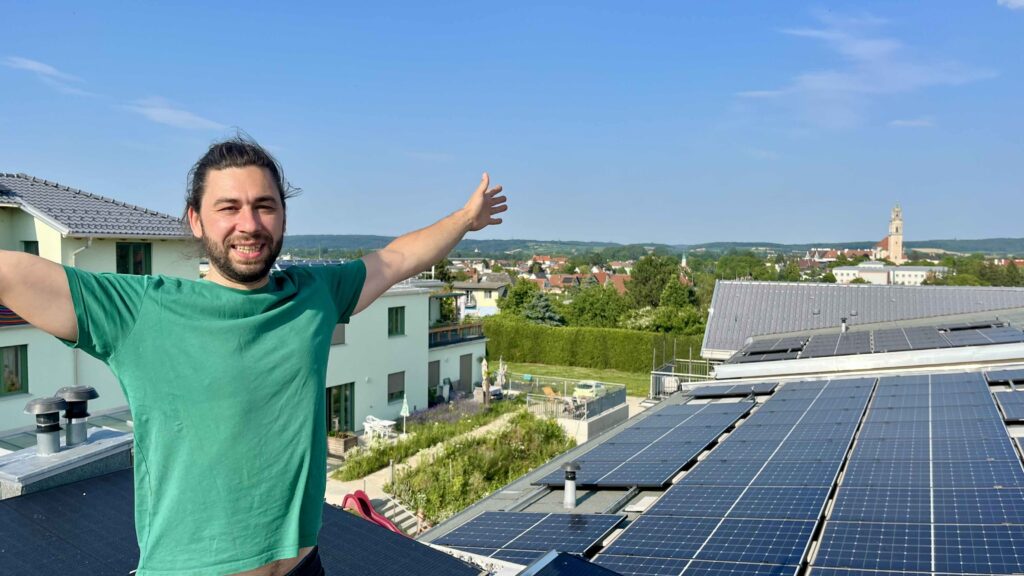 ein Mann steht auf einem Dach mit einer PV-Anlage. Er breitet die Arme aus.